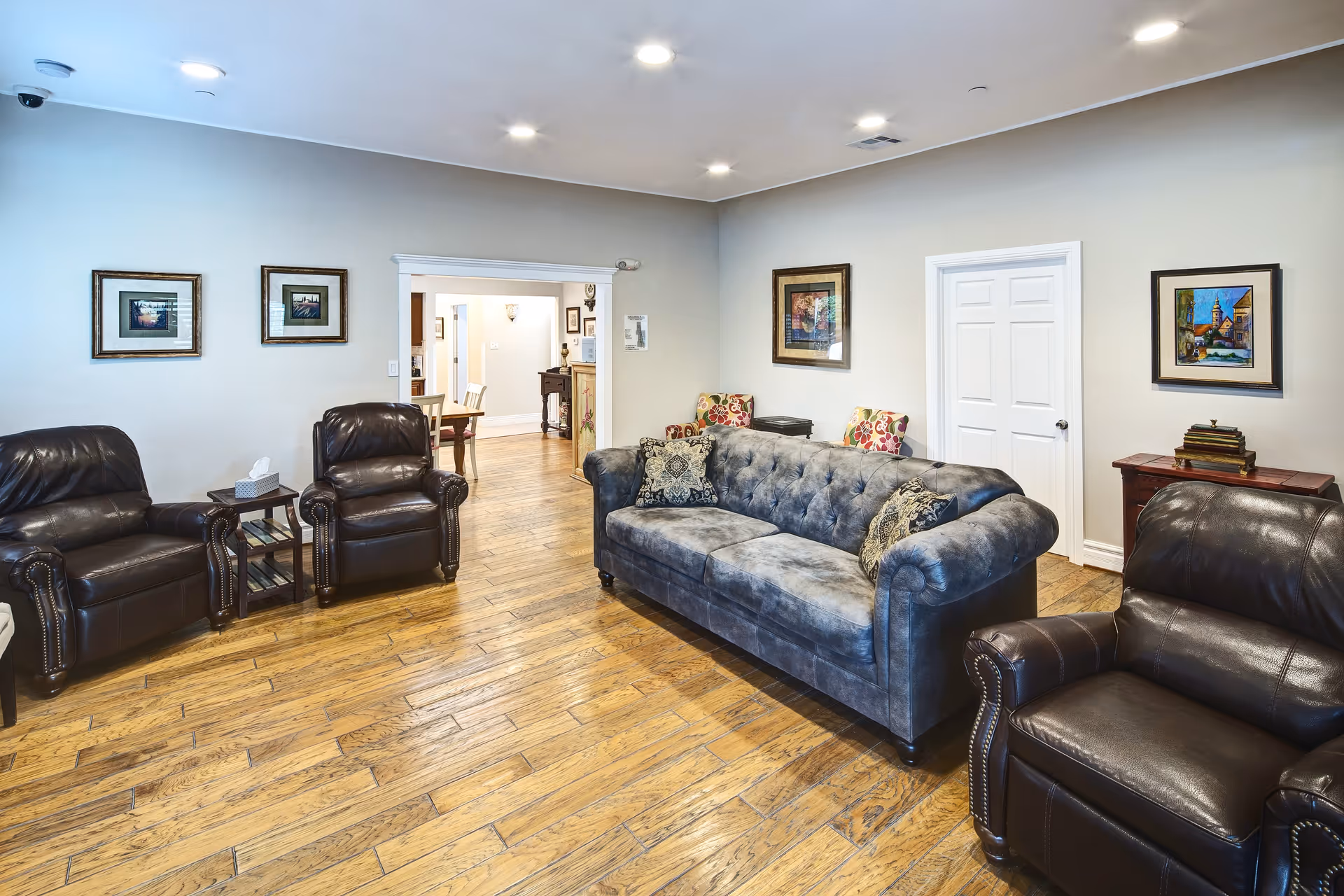 A cozy living room with wooden flooring, featuring a blue tufted sofa with patterned cushions, two dark brown leather armchairs, framed artwork on the walls, and a small side table with tissues. The room opens into a dining area with a wooden table and chairs.
