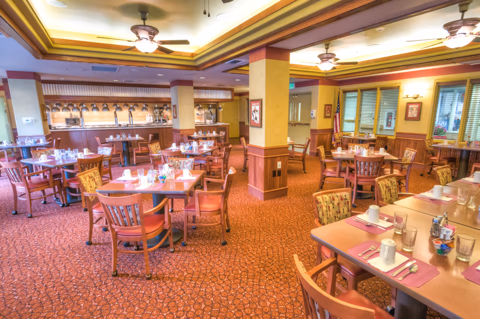 A spacious dining room with multiple tables and chairs arranged neatly. Each table is set with placemats, cups, glasses, and utensils. The room features warm lighting, ceiling fans, and large windows with blinds. The carpet has a patterned design, and there are columns and framed pictures on the walls.