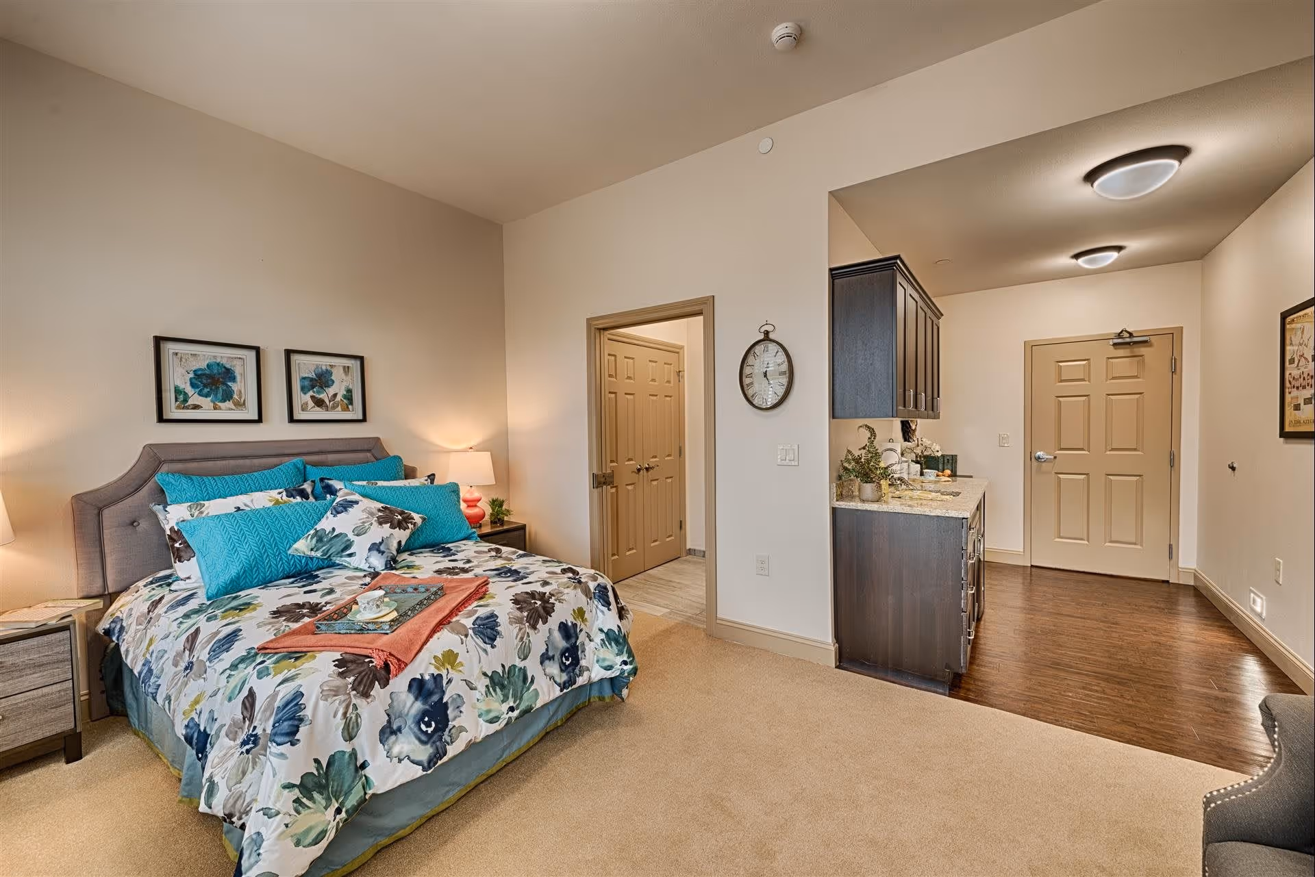 A cozy bedroom in a senior living facility with a bed featuring floral bedding and multiple blue pillows. There are two framed floral artworks above the bed, a nightstand with a lamp and a plant, and an open doorway leading to a bathroom. The room has beige walls and carpet, with a small kitchenette area visible near the entrance door.