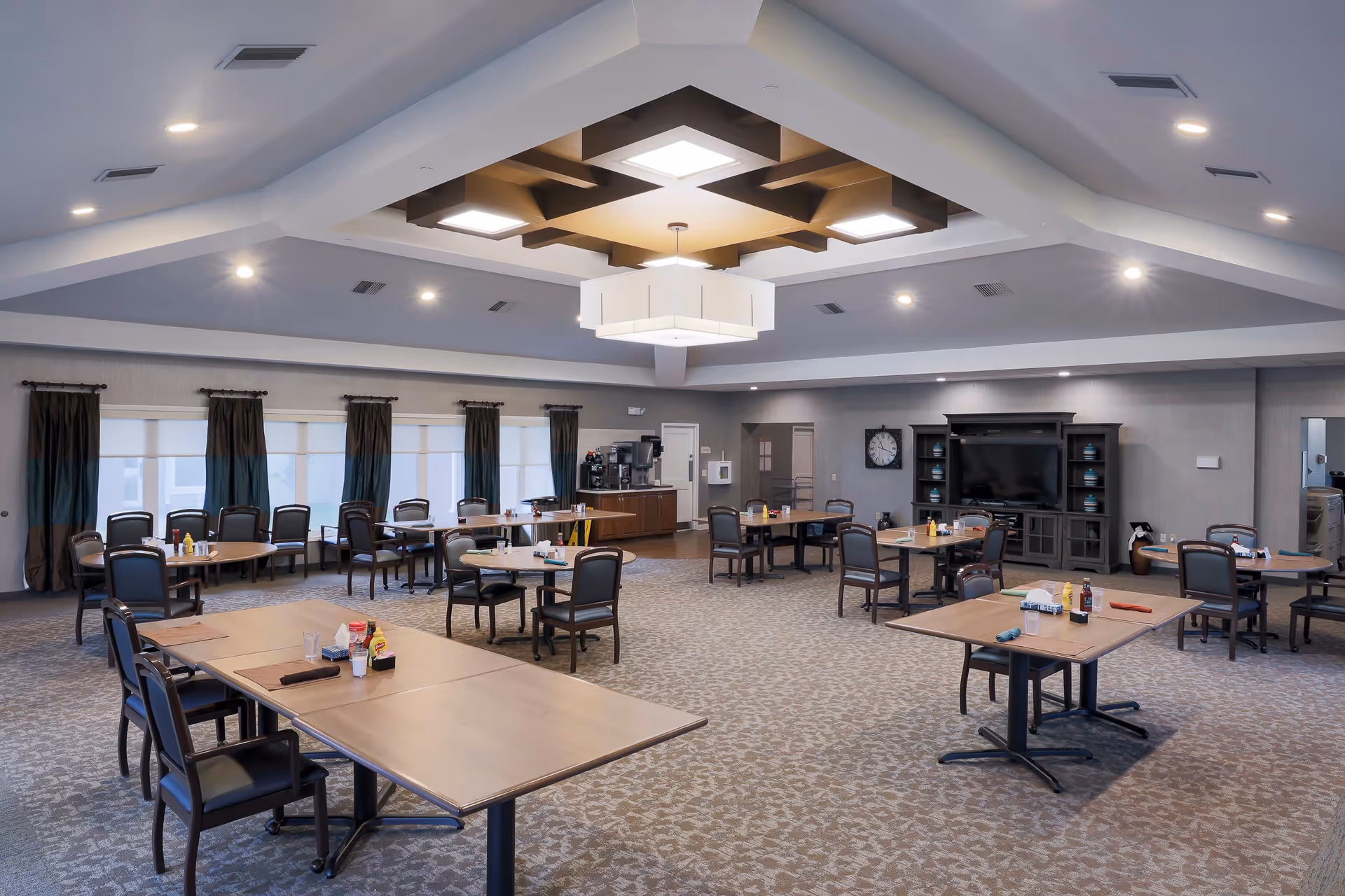 A spacious dining room with multiple round and rectangular tables, each surrounded by chairs. The tables are set with condiments and napkins. The room features a patterned carpet, large windows with dark curtains, a ceiling with recessed lighting and a central decorative light fixture. There is a large TV and cabinet on one wall, and a coffee station in the back corner.