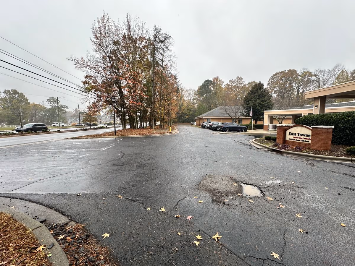 Wet driveway and parking area leading to the entrance of the East Towne facility with trees, parked cars, and a brick sign.