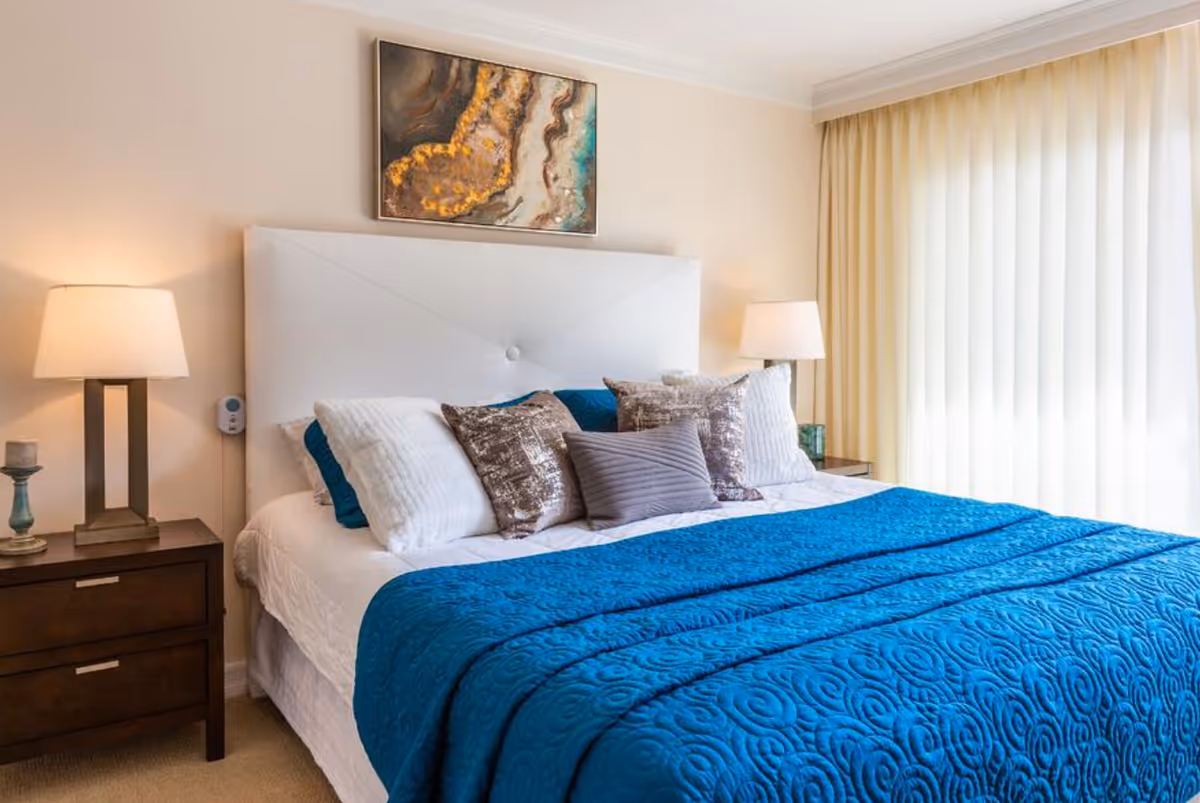 A neatly made bed with a white upholstered headboard, adorned with multiple pillows in white, brown, and gray colors, and covered with a blue quilted blanket. There are two bedside tables, each with a lamp, and a piece of abstract artwork hanging above the bed. Light-colored curtains cover a window on the right side of the room.