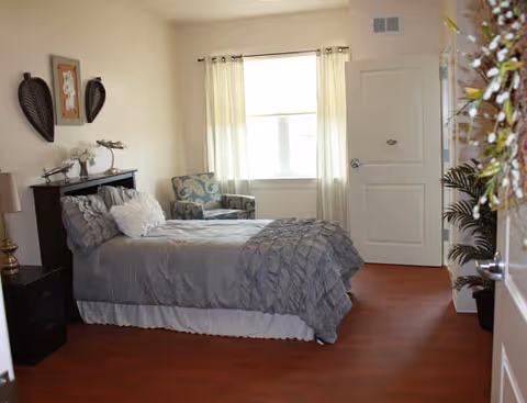 A bedroom with a neatly made bed covered in gray bedding, a dark wooden headboard, and a white bed skirt. There is a patterned armchair next to a window with light curtains. The room has wooden flooring, a nightstand with a lamp, decorative wall art above the bed, and a potted plant near the door.