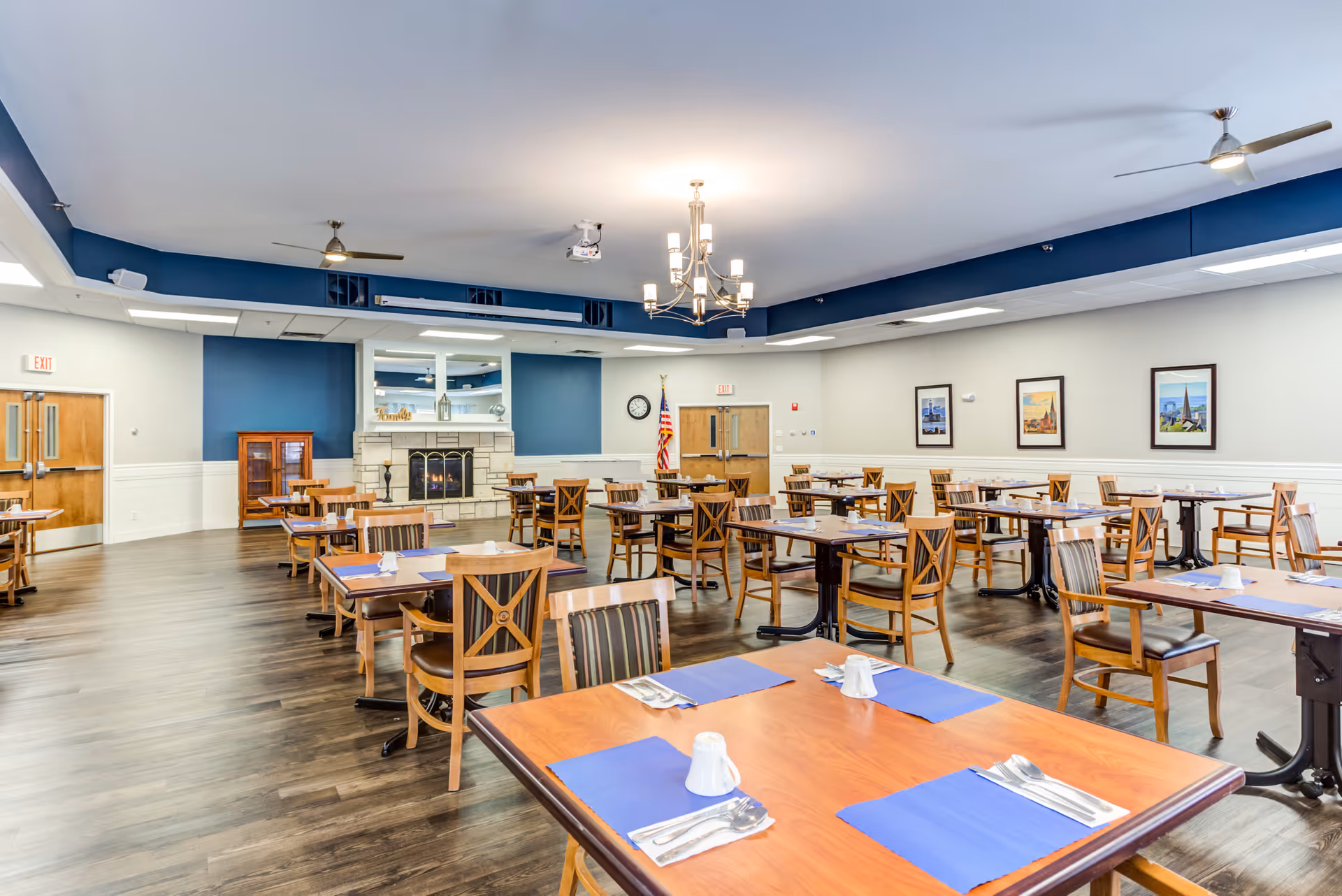 A spacious dining room with multiple wooden tables and chairs arranged neatly. Each table has blue placemats, white cups, and silverware. The room features a fireplace with a stone surround, a large mirror above it, and an American flag near double wooden doors. The walls are painted white with a blue accent wall behind the fireplace, and there are framed pictures hanging on the walls. The ceiling has modern light fixtures and ceiling fans.