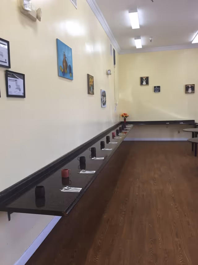 A long narrow dining area with a wall-mounted black countertop set with black and red mugs and silverware. The walls are painted light yellow and decorated with framed pictures and certificates. The floor is wooden, and there are ceiling lights illuminating the space.