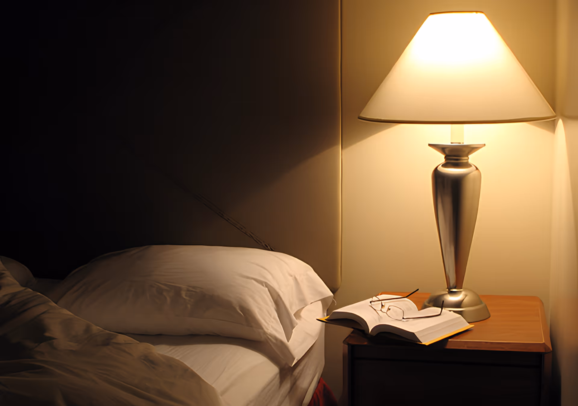 A bedside table with a silver lamp emitting warm light, an open book, and a pair of reading glasses resting on the book. Next to the table is a bed with white pillows and bedding, partially visible in a dimly lit room.