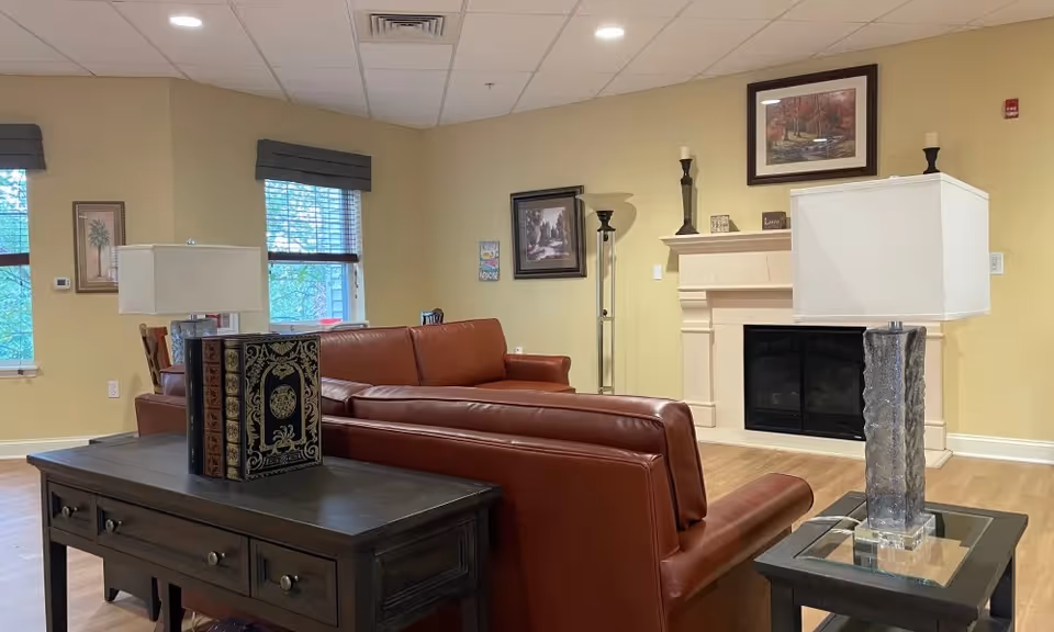 A cozy living room area with two brown leather sofas facing a white fireplace. There are two table lamps with white shades on dark wooden tables, decorative books on one table, and framed artwork on the beige walls. The room has wooden flooring and windows with gray valances.