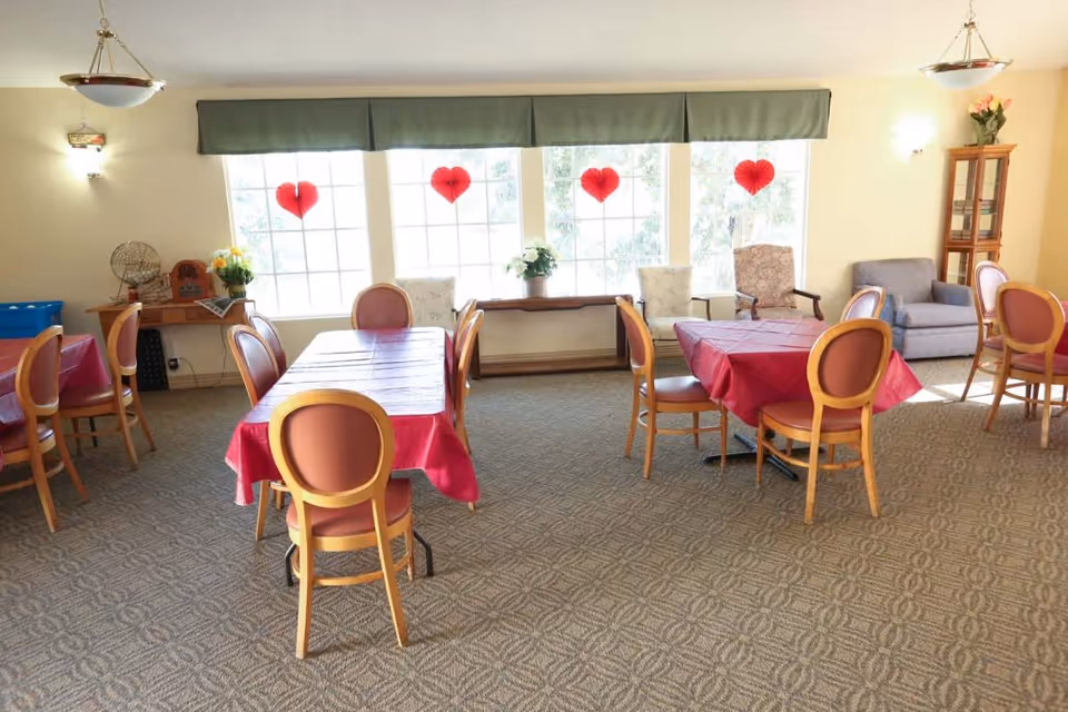 A bright dining room with several tables covered in red tablecloths and surrounded by wooden chairs with red cushions. Large windows with green valances let in natural light and have red heart decorations hanging. There are a few armchairs and a small wooden cabinet with flowers in the background.