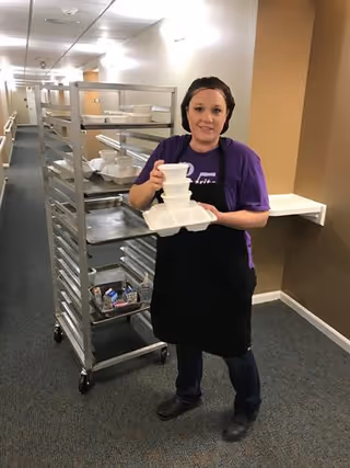 A woman wearing a purple shirt and black apron stands in a hallway holding a stack of white food trays. Behind her is a metal rack with multiple shelves holding more food trays and cartons of milk.