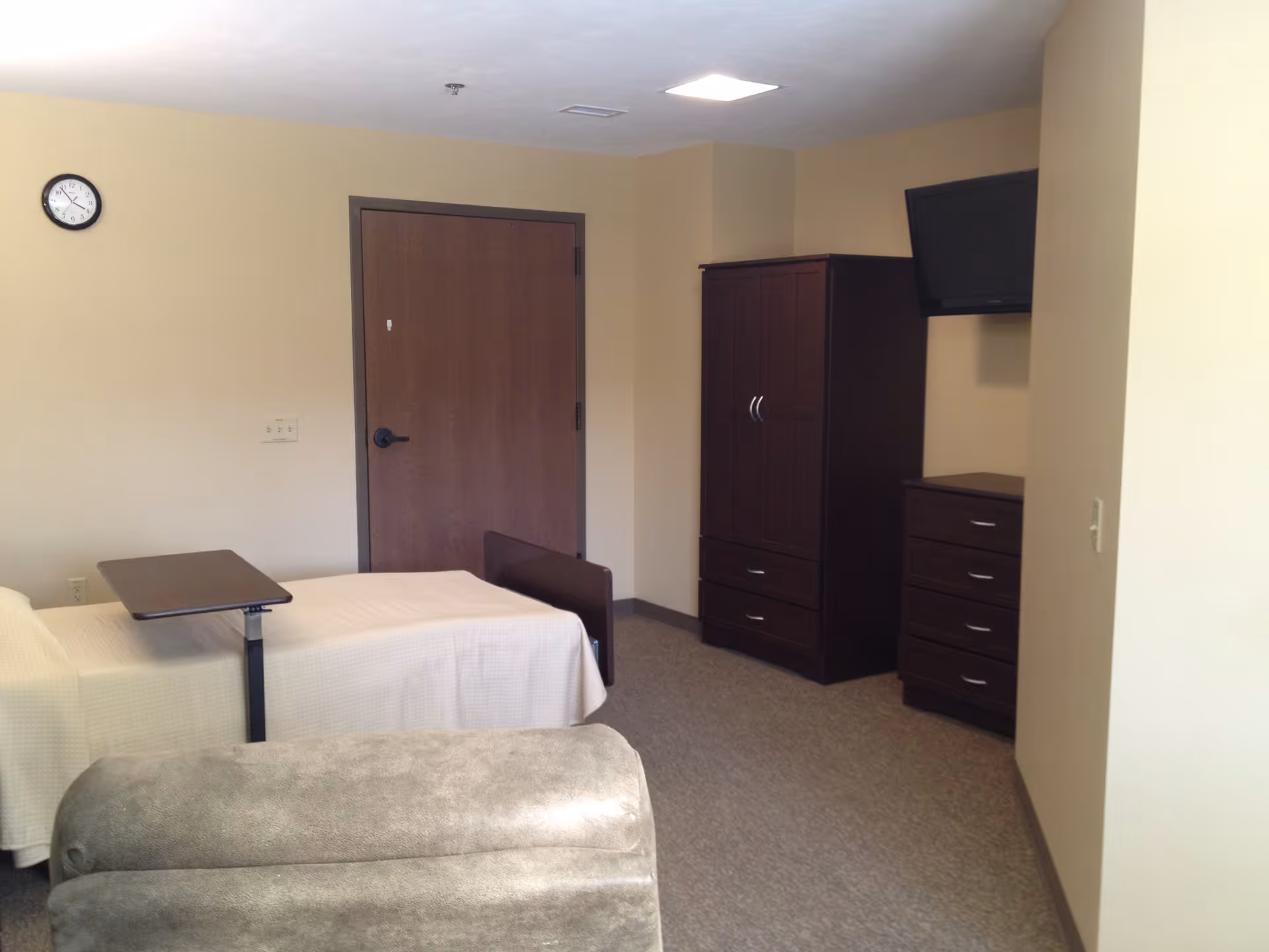 Furnished bedroom with a single bed and overbed table, recliner in the foreground, and a wardrobe, dresser and wall-mounted TV near a closed door.
