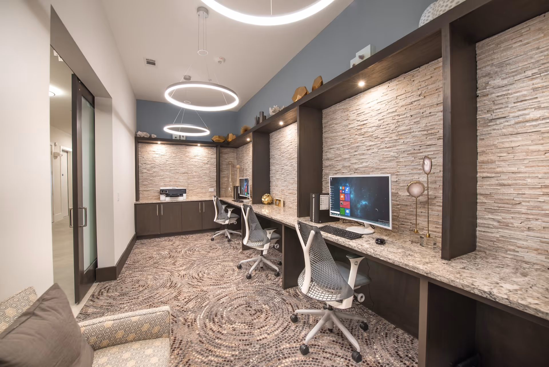 Modern computer workstation area with three ergonomic chairs and desktop computers on a long granite countertop. The wall behind the desks features textured stone panels with decorative items on shelves above. Circular pendant lights hang from the ceiling, and a patterned carpet covers the floor. A glass door is visible on the left side.