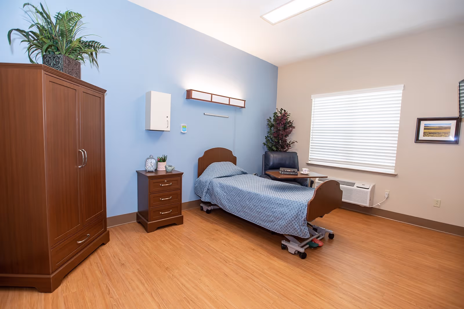 A clean resident bedroom with a single adjustable bed, wooden wardrobe and nightstand, a chair, and a window with blinds.