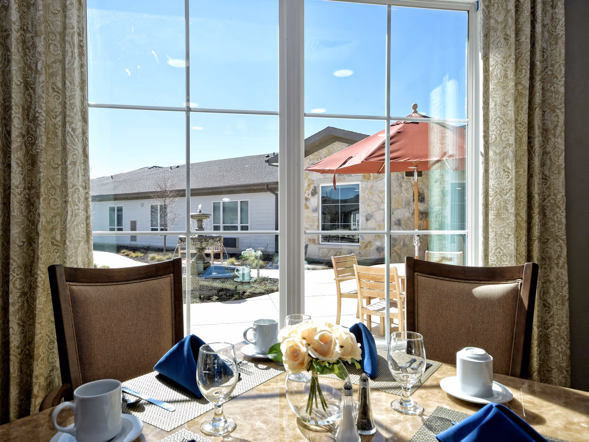 A dining table set for four with glasses, cups, silverware, and blue napkins in front of a large window. Outside the window, there is a patio area with wooden chairs, a table, a red umbrella, and a stone building with a fountain in the background.