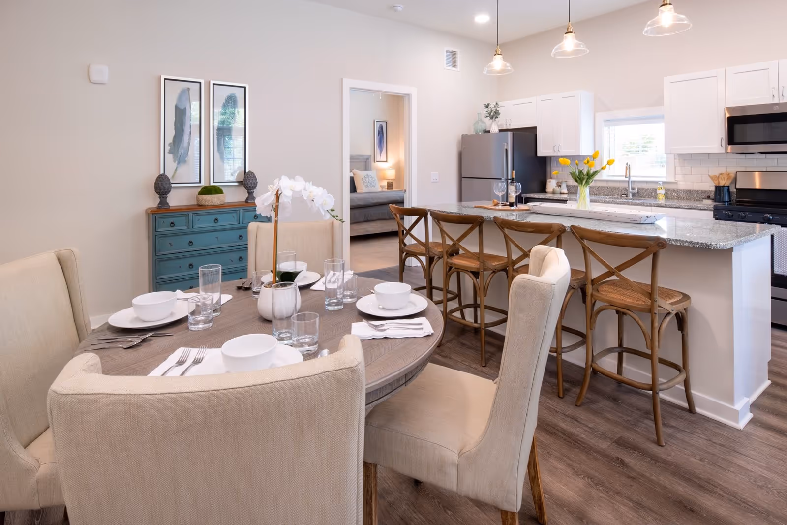 A bright and modern dining area with a round wooden table set for four with white dishes, glasses, and silverware. Beige upholstered chairs surround the table. In the background, there is a kitchen with white cabinets, a granite countertop island with four wooden bar stools, stainless steel appliances, and a vase of yellow tulips. A doorway leads to a bedroom with a bed and framed artwork on the wall. The room has wood flooring and neutral-colored walls.