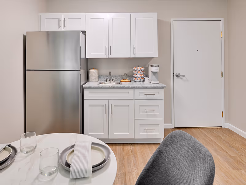 A small kitchen area with white cabinets, a stainless steel refrigerator, a countertop with a sink, a coffee maker, and a container. In the foreground, there is a round dining table set with plates, glasses, and napkins, and a gray chair is partially visible. The floor is wooden, and there is a closed white door on the right side.