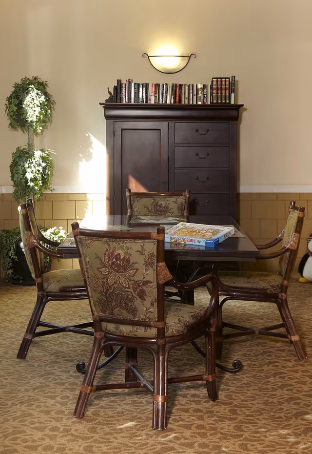 A cozy interior room featuring a square wooden table with a glass top surrounded by four cushioned chairs with floral upholstery. On the table, there is a puzzle box. Behind the table is a dark wooden cabinet with books on top and a wall-mounted light fixture above it. To the left, there is a tall green potted plant. The room has beige walls and patterned carpet flooring.