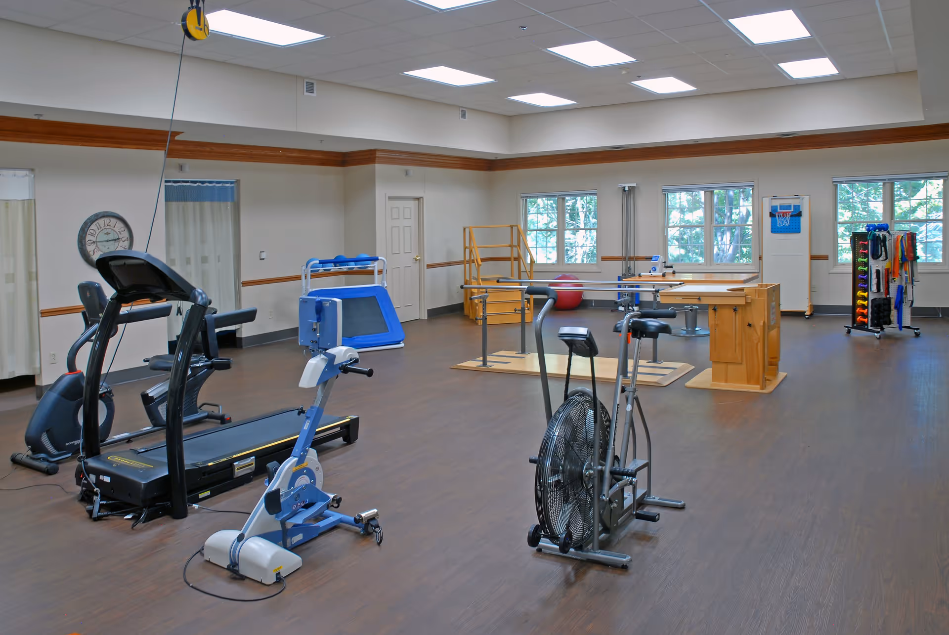 A spacious senior living facility exercise room with various fitness equipment including a treadmill, stationary bike, and other rehabilitation apparatus. The room has large windows letting in natural light, wooden flooring, and light-colored walls with wood trim. There are also therapy tools and a small basketball hoop on the wall.