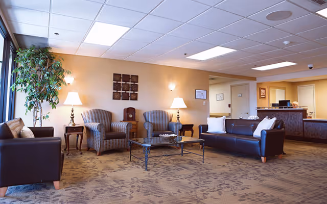 A spacious assisted living facility common area with a brown leather sofa, two patterned armchairs, a glass coffee table, side tables with lamps, a large potted plant near a window, and a reception desk in the background.