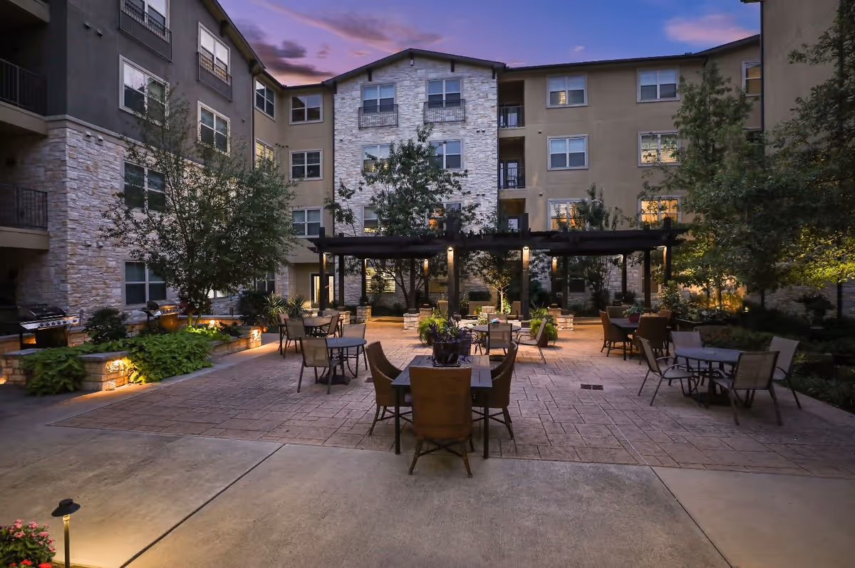 Outdoor courtyard area of a senior living facility at dusk with multiple tables and chairs arranged on a paved surface, surrounded by trees and plants. The building in the background has multiple windows and balconies, with warm lighting inside. There is a pergola structure with lights and some built-in grills on the left side.