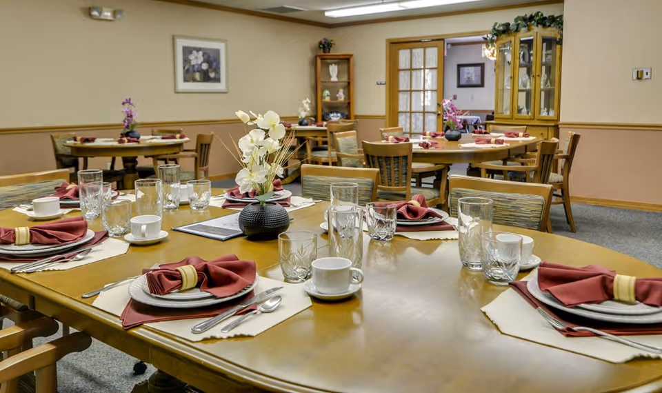 A dining room in a senior living facility with multiple round wooden tables set with plates, cups, glasses, silverware, and folded red napkins. Each table has a small floral centerpiece. The room has beige walls with wooden trim, framed artwork, and wooden chairs with patterned cushions. There are cabinets and shelves with decorative items in the background.