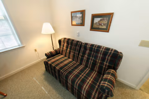 A small living area with a plaid-patterned sofa against a white wall. Two framed pictures hang above the sofa. A floor lamp is positioned next to the sofa near a window with blinds. The floor is carpeted.