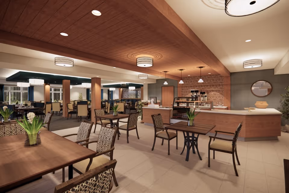 A spacious and well-lit dining area in a senior living facility featuring multiple wooden tables and chairs with patterned upholstery. The room has a modern design with a wooden ceiling accent, pendant lights, and a service counter with coffee machines and display cases. There are decorative plants on the tables and a round mirror on the wall behind the counter.