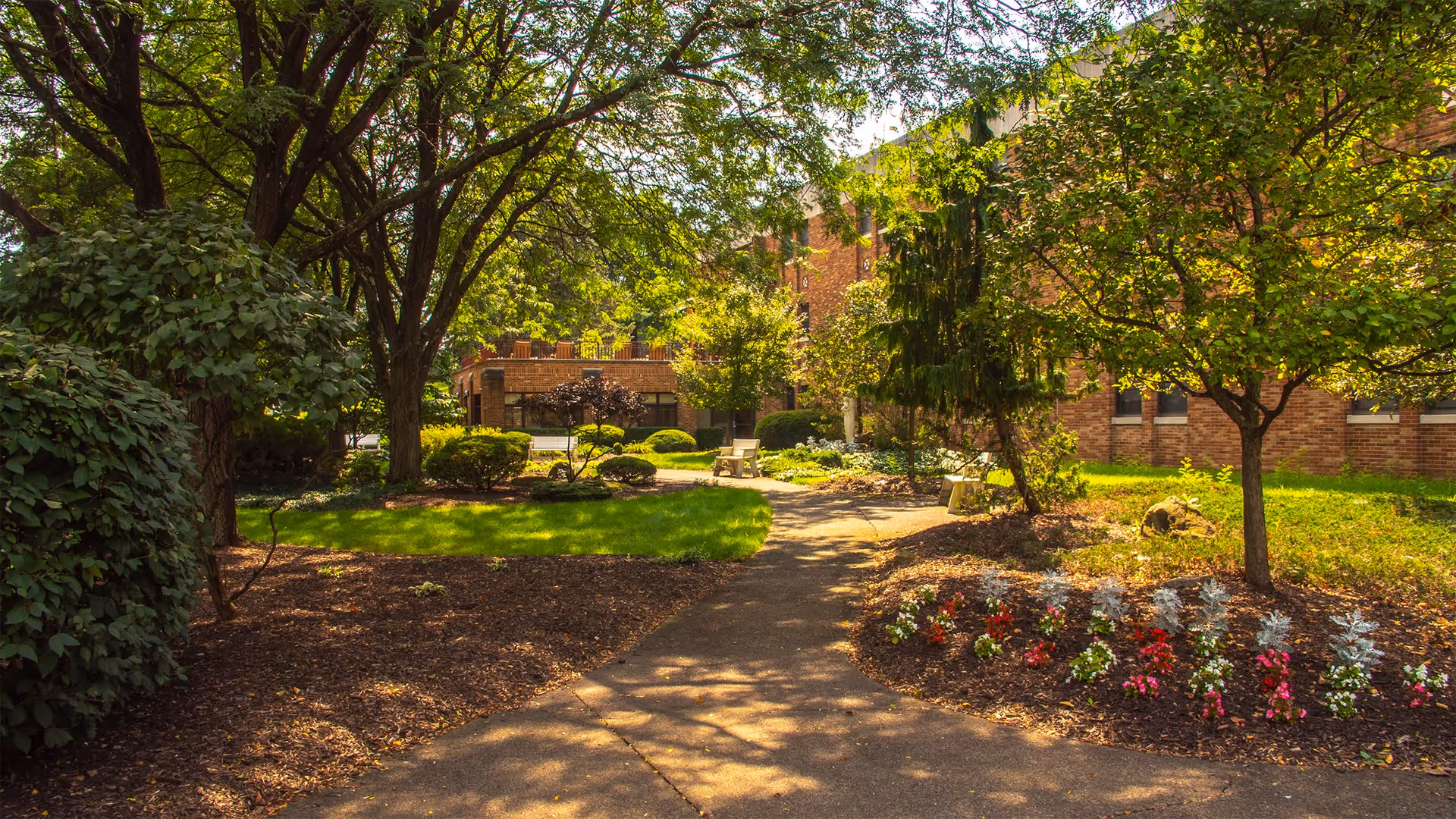A sunny outdoor garden area with a paved walkway surrounded by green grass, trees, shrubs, and colorful flower beds. There are benches along the path and a brick building in the background.