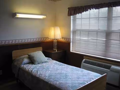 A single bed in a small bedroom with a lamp, window blinds, and air conditioner.