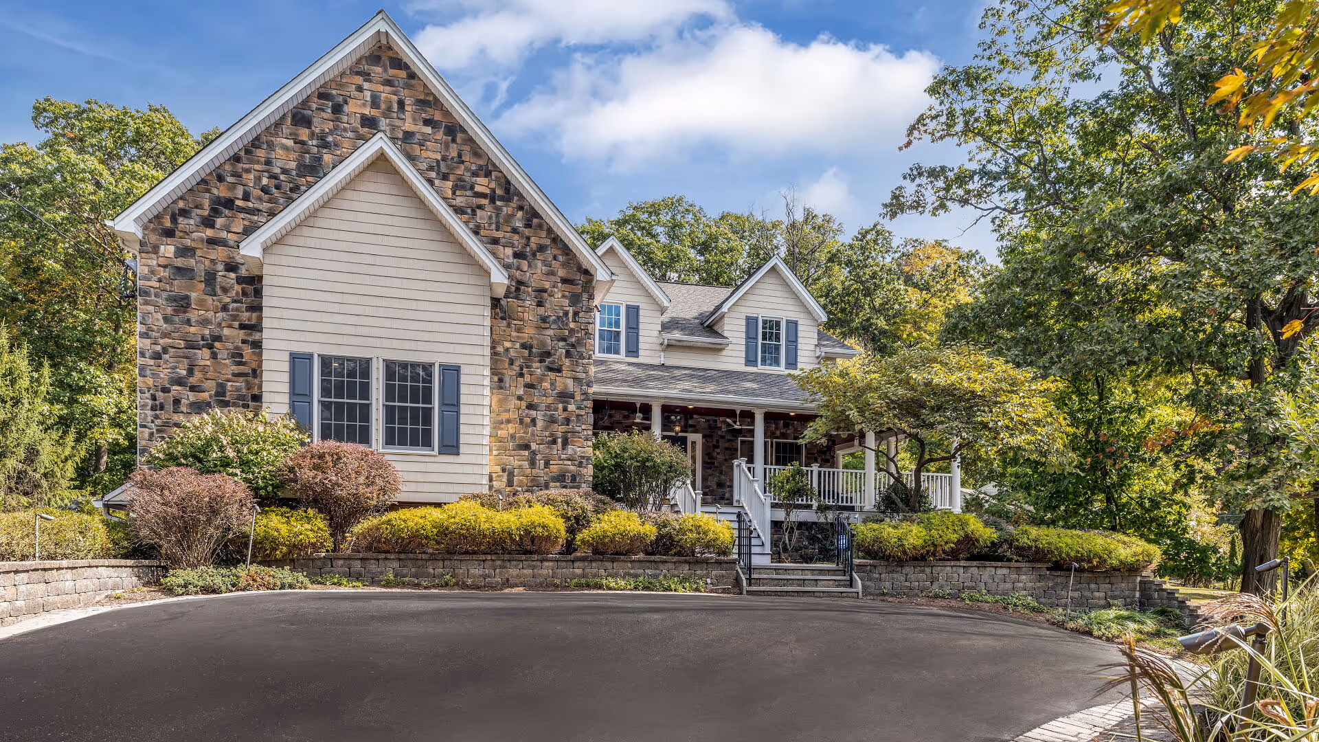 Front exterior view of a senior living facility named Clover Hill Senior Living, featuring a stone and siding facade, multiple windows, a covered porch with white railings, surrounded by landscaped bushes and trees under a partly cloudy sky.
