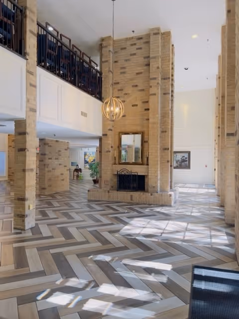 Spacious interior area of a senior living facility with tall brick pillars, a central brick fireplace with a mirror above it, and a modern hanging light fixture. The floor features a herringbone pattern with light and dark wood tones. There is a balcony with dark railing on the upper level and large windows allowing natural light to fill the space.