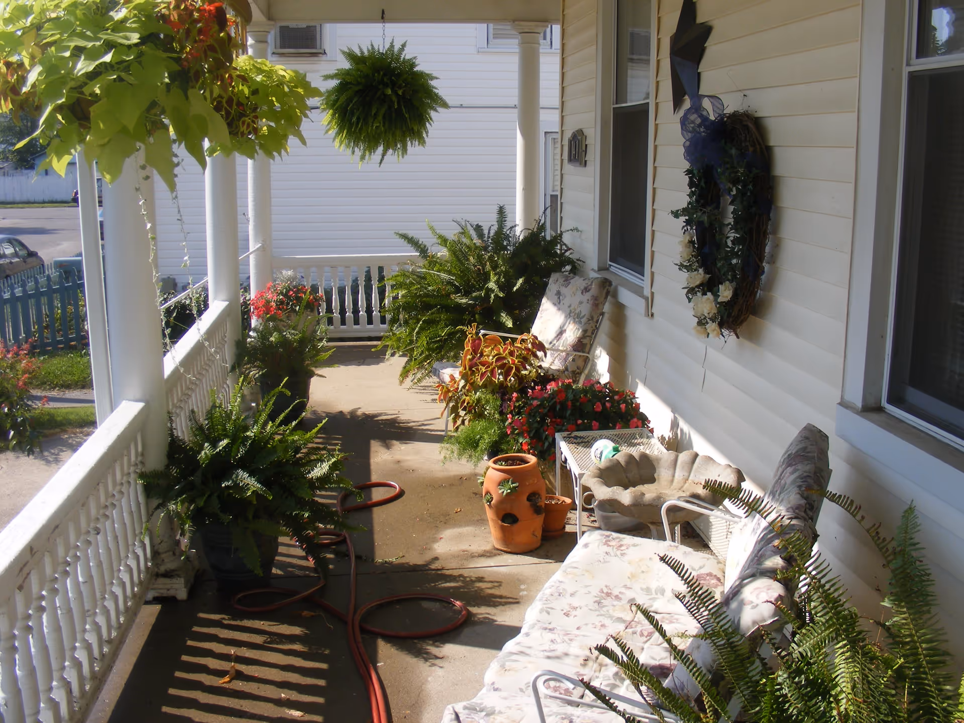 A sunny porch area with white railings and columns, decorated with various potted plants and hanging ferns. There are cushioned chairs and a small table with a decorative birdbath. A wreath with white flowers and a blue ribbon hangs on the wall of the building.