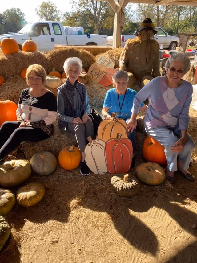 Four elderly women sitting on hay bales surrounded by pumpkins and decorative wooden pumpkins under a wooden shelter. Behind them is a scarecrow and a white pickup truck parked outside.