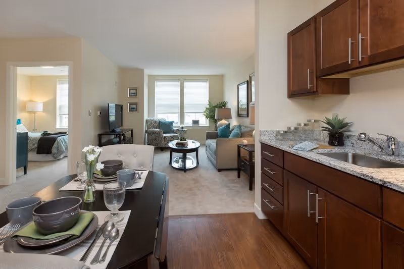 View of a modern apartment interior showing a small kitchen area with wooden cabinets and granite countertop on the right, a dining table set for two with dishes and glasses in the foreground, and a living room with a sofa, armchair, coffee table, and large window in the background. A bedroom with a bed and lamp is visible through an open doorway on the left.