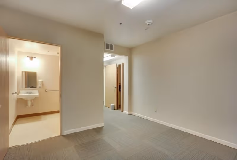 An empty interior room with beige walls and carpeted floor, showing an open doorway leading to a small bathroom with a sink, mirror, and light fixture above it. Another open doorway leads to a hallway with a partially visible door and a small trash can on the floor.
