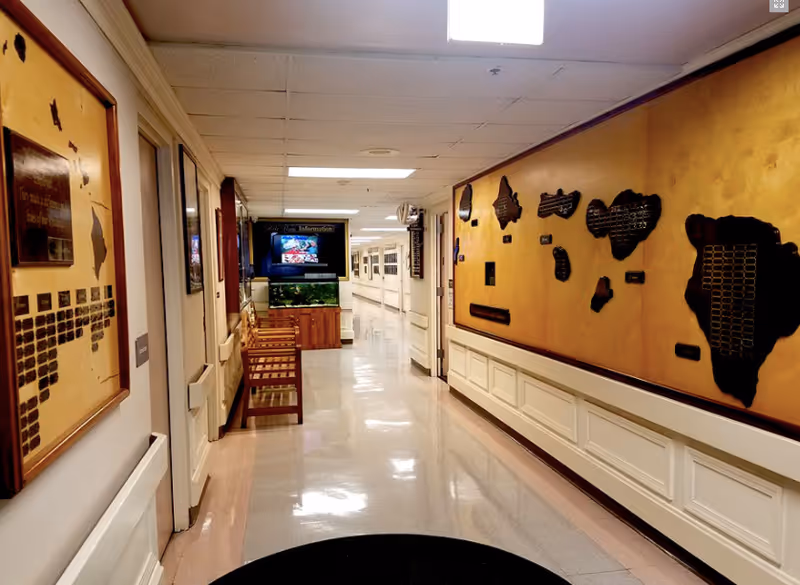 A clean, well-lit hallway in a senior living facility with polished floors, wooden chairs along the left wall, and large decorative plaques on the right wall. At the end of the hallway, there is a television mounted above a fish tank displaying information.