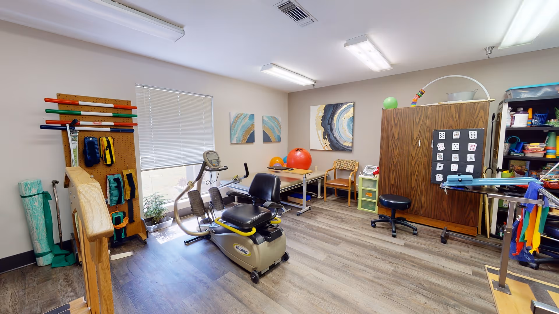 A physical therapy or rehabilitation room with exercise equipment including a stationary recumbent bike, parallel bars, therapy balls, and various therapy tools organized on shelves and a pegboard. The room has wood flooring, neutral-colored walls, two abstract paintings, and a window with blinds.