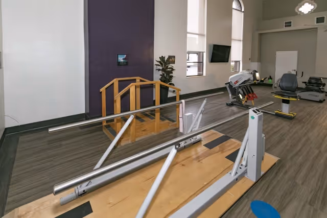 Interior view of a rehabilitation or physical therapy room with parallel bars for walking exercises, a small wooden ramp with handrails, exercise equipment including a recumbent bike and other fitness machines, large windows letting in natural light, and a plant against a purple accent wall.
