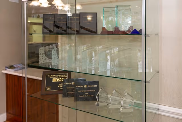 Glass display case with multiple awards and plaques inside, including recognitions from the California Association of Health Facilities and Ensign Services, located in a room with beige walls and wooden cabinetry.