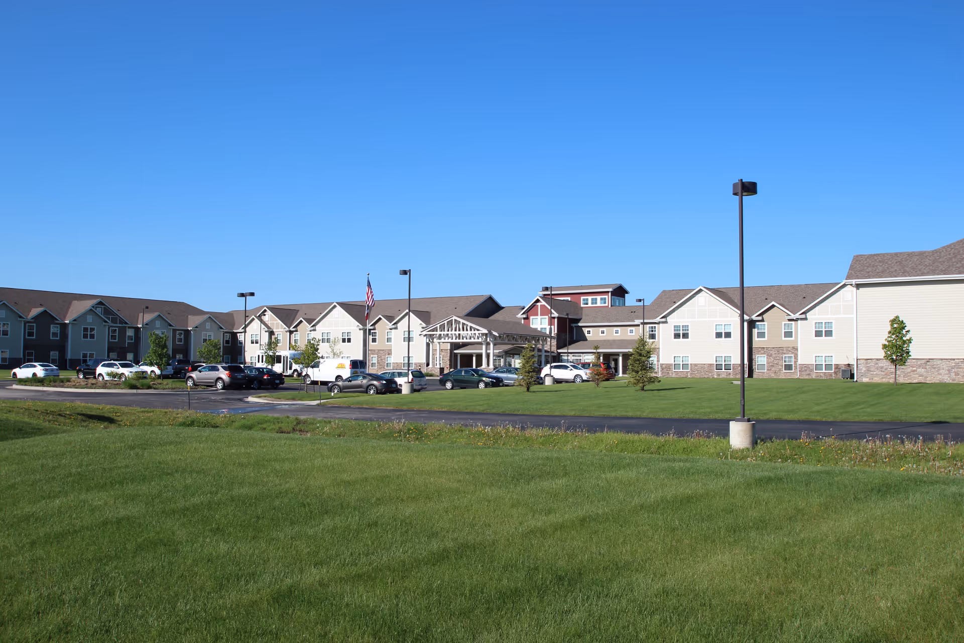 Wide exterior view of Kennybrook Village senior living facility under a clear blue sky, showing a large, multi-section building with beige and red siding, multiple windows, a covered entrance, a parking lot with several cars, green lawns, and a few small trees.