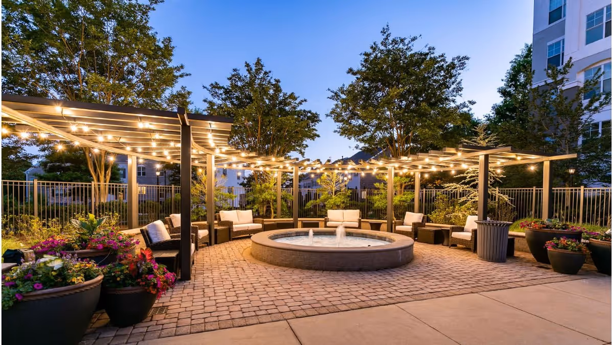 Outdoor seating area at dusk with a circular water fountain in the center, surrounded by cushioned wicker chairs under pergolas adorned with string lights. Large potted plants and trees are visible around the fenced area.