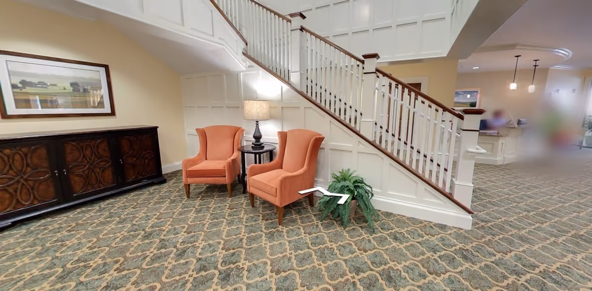 Interior view of a senior living facility lobby area with a staircase. Two orange armchairs are placed beside a small round table with a lamp. A large wooden cabinet with decorative doors is against the wall, and a framed landscape painting hangs above it. The floor is carpeted with a patterned design, and there is a potted plant near the staircase. In the background, a reception desk and blurred figures are visible.