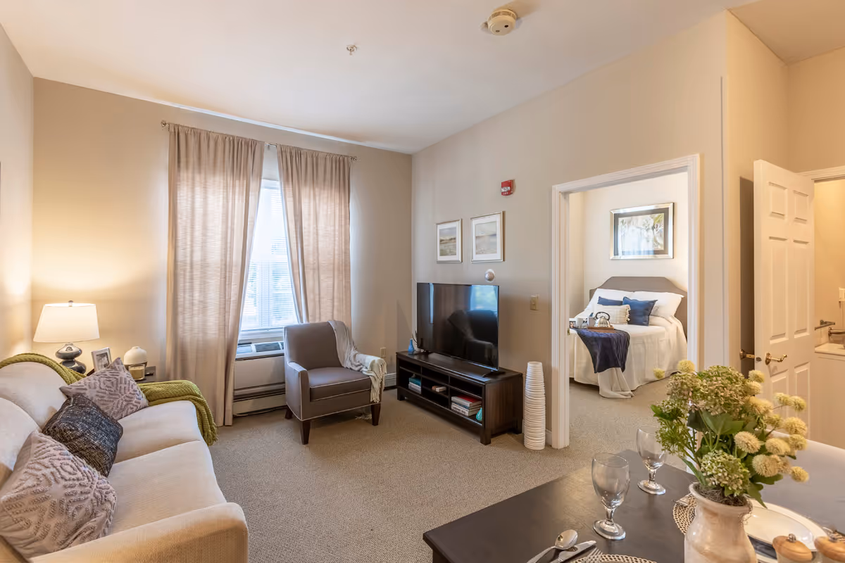 A cozy living room with a beige sofa adorned with pillows and a green throw blanket, a gray armchair, and a flat-screen TV on a dark wooden stand. The room has beige walls and carpet, with a window covered by light beige curtains. Through an open doorway, a bedroom with a neatly made bed featuring white and blue bedding is visible. A dining table with place settings and a vase of flowers is partially visible in the foreground.