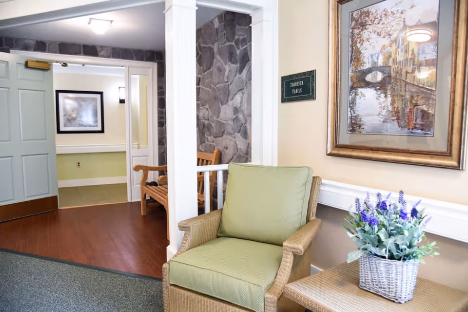 A cozy seating area in a senior living facility featuring a green cushioned wicker chair next to a wicker table with a potted plant with purple flowers. Behind the chair is a framed painting of a canal scene with buildings and trees. The wall has a small sign that reads 'Zinnesta Trails.' In the background, there is a wooden bench against a stone wall and an open light blue door leading to another room with a framed picture on the wall.