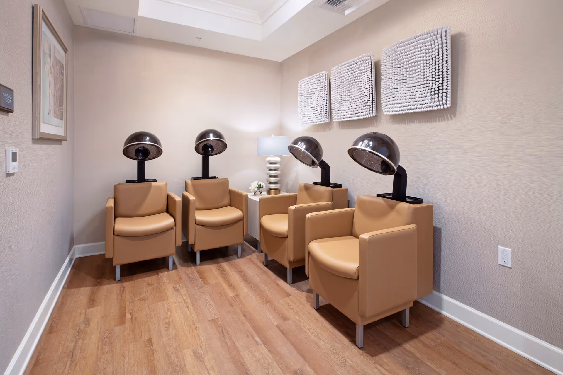 A small room with four tan leather chairs each equipped with a black hair dryer hood. The room has light beige walls, wooden flooring, a white side table with a modern lamp and a small flower arrangement, and three textured white wall art pieces. A sign on the left wall reads 'Massage Room'.