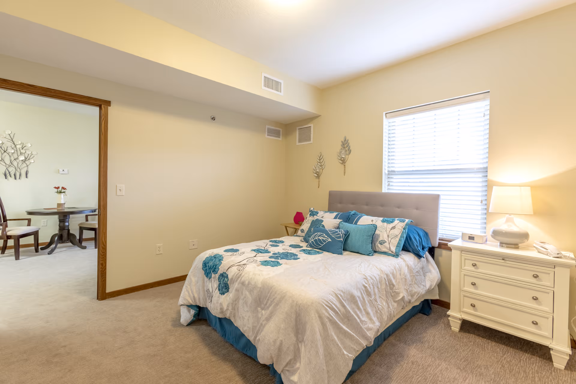 Bright bedroom with a bed in blue-and-white bedding, a nightstand and lamp by a window, and a doorway opening to a small dining/sitting area.