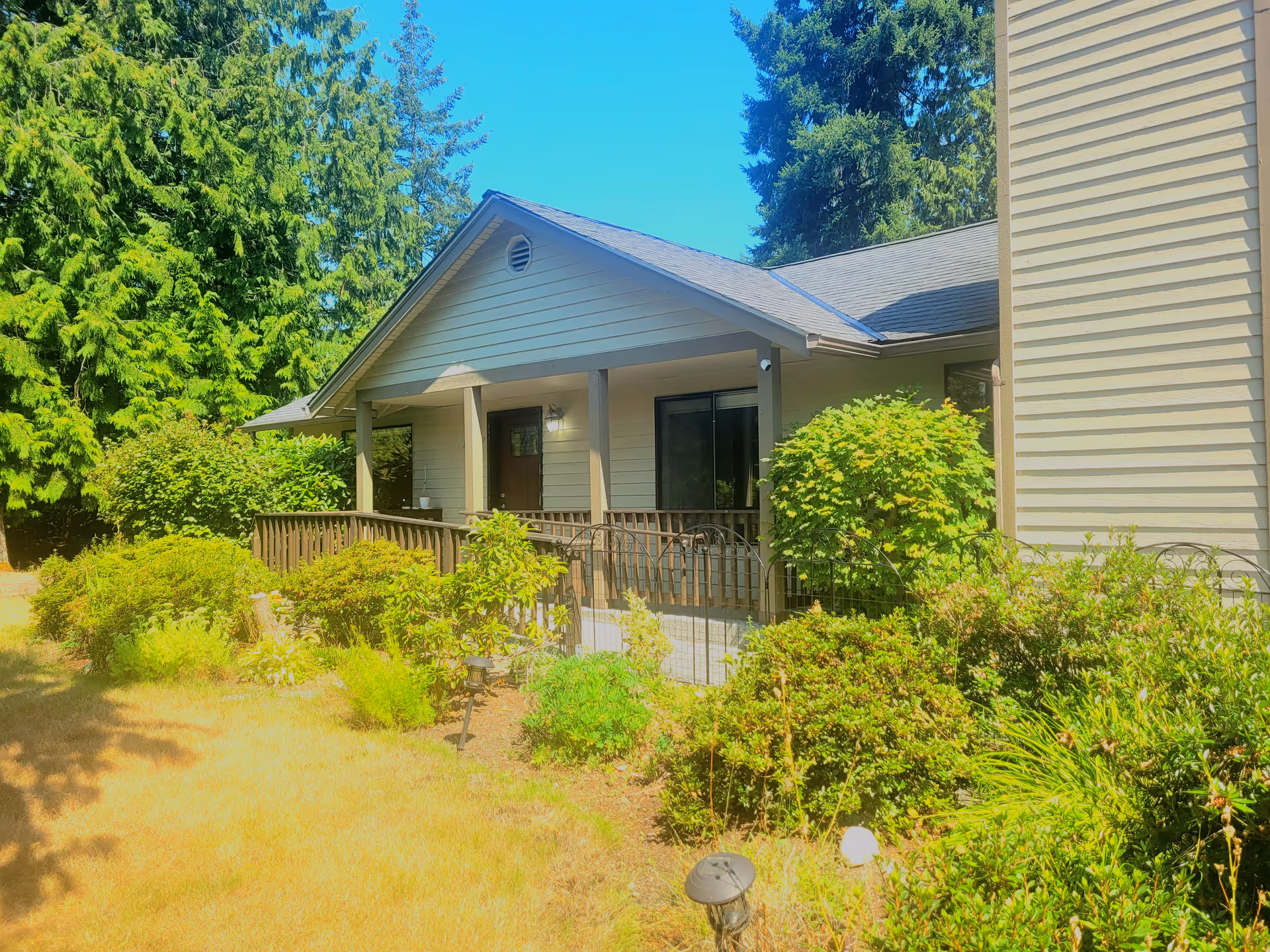 Exterior view of a single-story building with beige siding and a covered porch. The porch has a wooden railing and is surrounded by green bushes and plants. Tall evergreen trees are visible in the background under a clear blue sky.