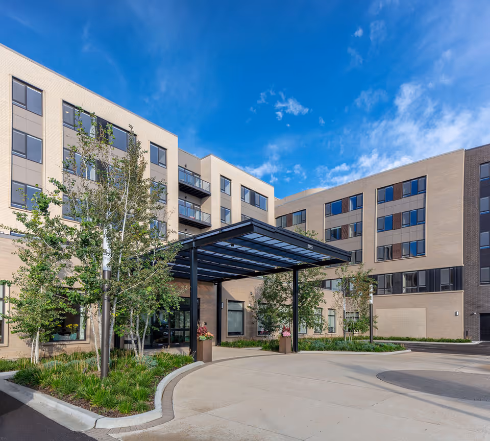 Entrance and facade of a modern multi-story senior living building with a covered drop-off canopy, landscaping, and a blue sky.