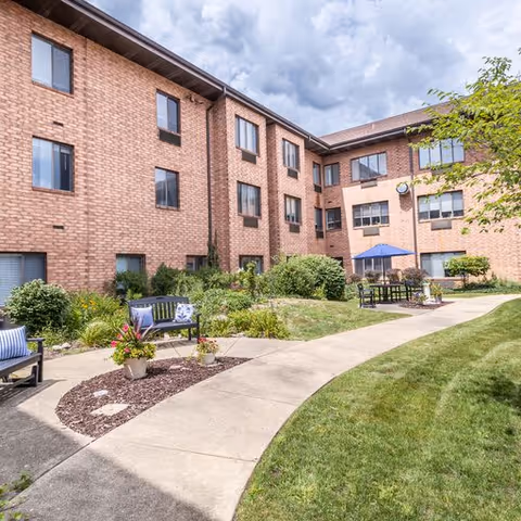 Outdoor courtyard area of a senior living facility with a curved concrete walkway, green grass, landscaped flower beds, benches with cushions, and a brick building with multiple windows in the background under a cloudy sky.