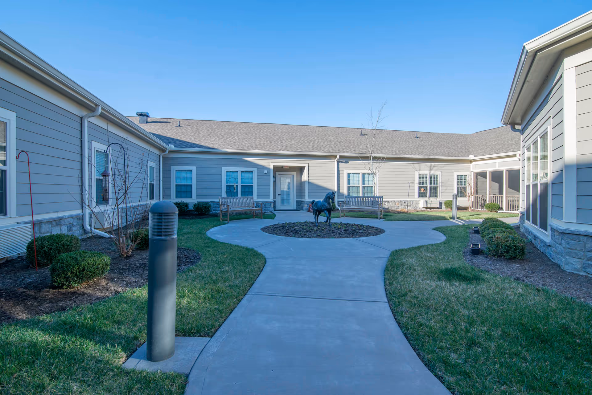 A paved courtyard walkway leading to a central circular planting with a horse sculpture, surrounded by lawns, benches and single-story beige buildings under a clear sky.
