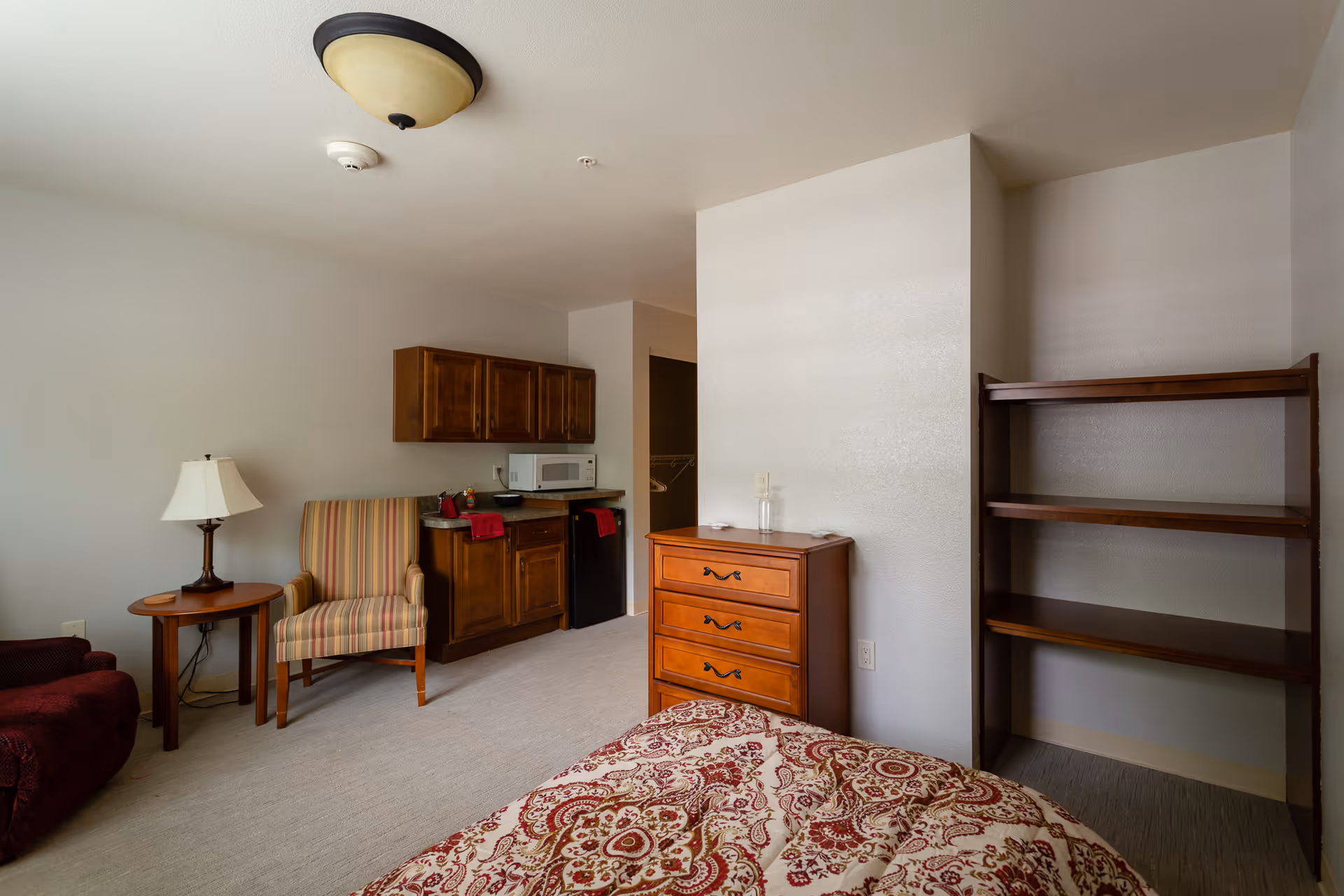 Interior view of a senior living facility room featuring a bed with a patterned bedspread, a wooden dresser, an empty wooden shelving unit, a striped armchair next to a small round table with a lamp, and a kitchenette area with wooden cabinets, a microwave, and a mini fridge.